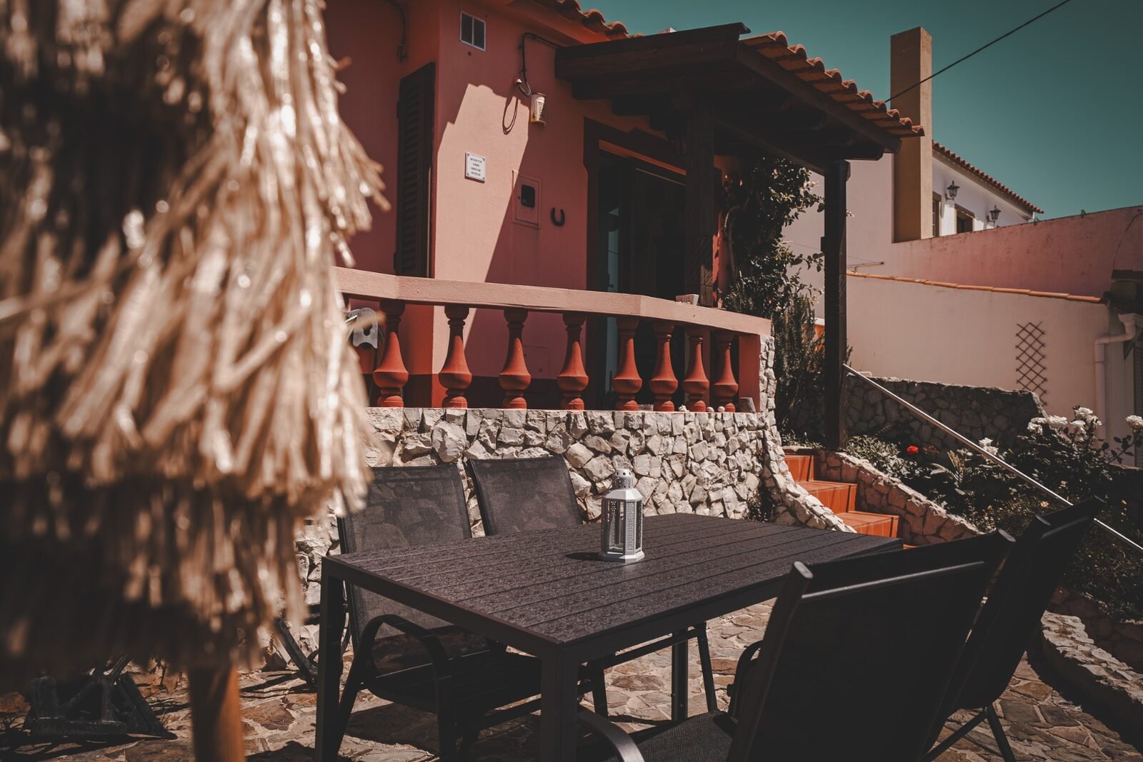 The warm coral façade and stone terrace of Casa Budens, with outdoor dining and a straw umbrella in the Algarve sun